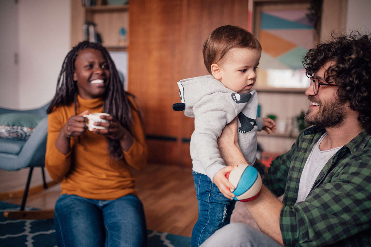 A foster dad plays and cuddles with his toddler son while his wife smiles and looks on in the background.