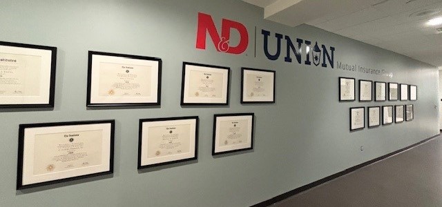 Framed professional designation certificates displayed along a hallway wall at ND & Union Mutual Insurance Group.