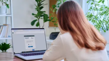 Woman sitting at a desk using a laptop displaying an online proctoring system