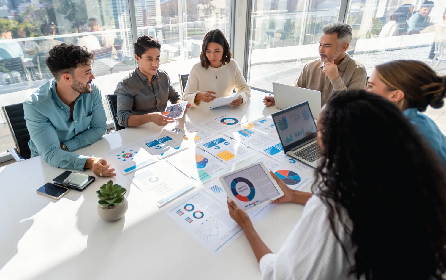 Group of professionals in a modern office sitting around a table covered with charts and reports, collaborating on data and business strategy.