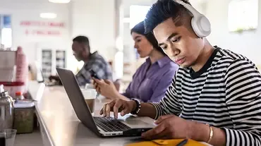 Student multitasks with headphones on, navigating both laptop and phone in a modern, collaborative workspace.