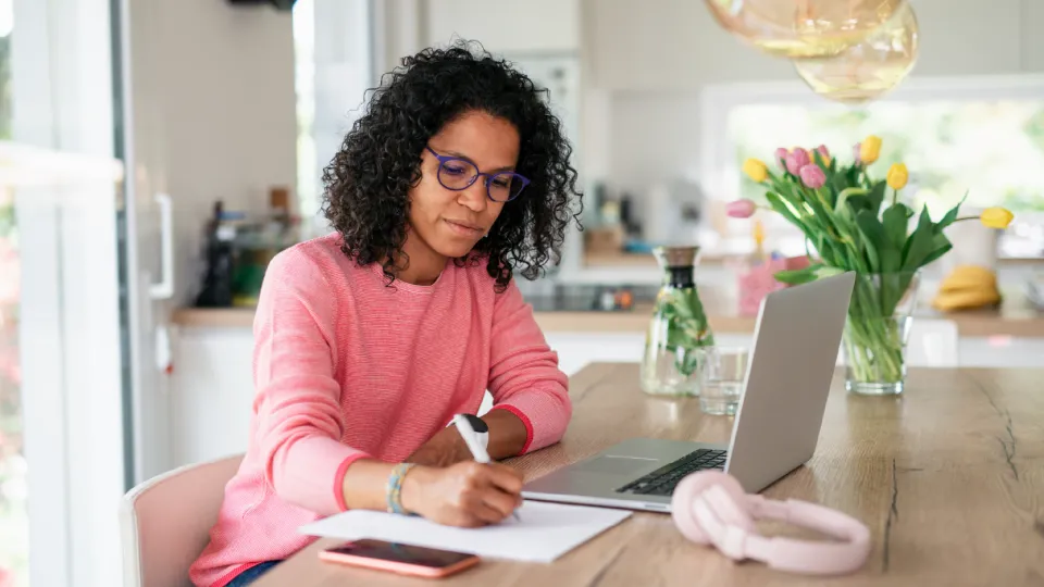 Woman with pink sweater writing on a notepad in front of a laptop while sitting on a kitchen table.