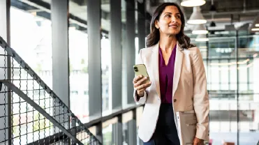 Confident businesswoman holding a phone and laptop walks through a modern office space, appearing focused and ready for the workday.