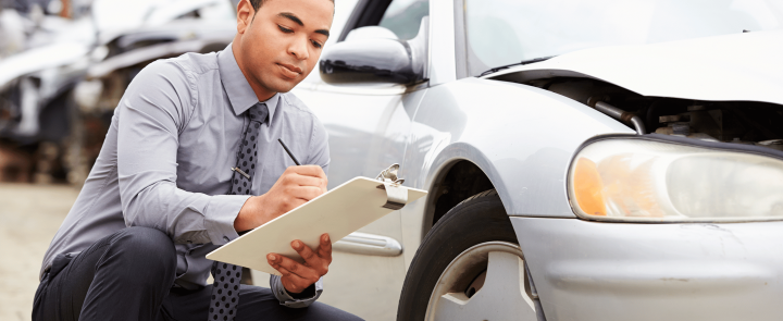 Insurance adjuster kneeling next to a damaged car, writing notes on a clipboard during a vehicle inspection.