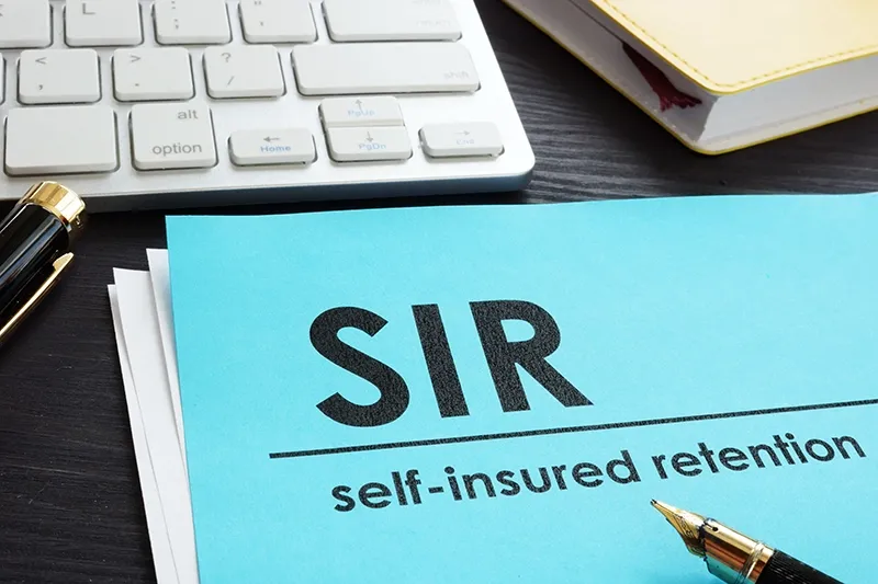 A blue folder labeled "SIR self-insured retentions" sits on a desk with a pen next to a computer keyboard.