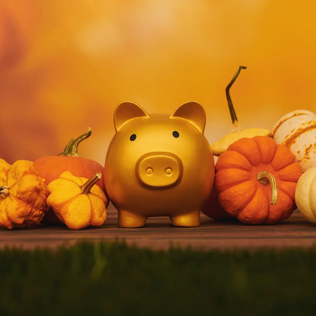 A golden piggy banks sits in front of an autumnal orange background surrounded by decorative fall gords.