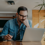 Man with glasses sitting at the table looking at laptop and taking notes.