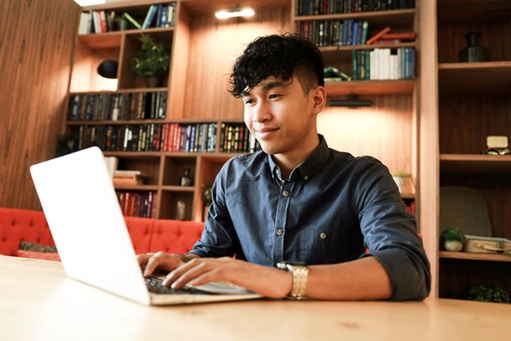 A young man in a blue shirt works on a laptop in front of a bookcase.
