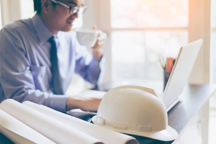 A construction foreman sits at a computer with rolled building plans and a hardhat in the foreground