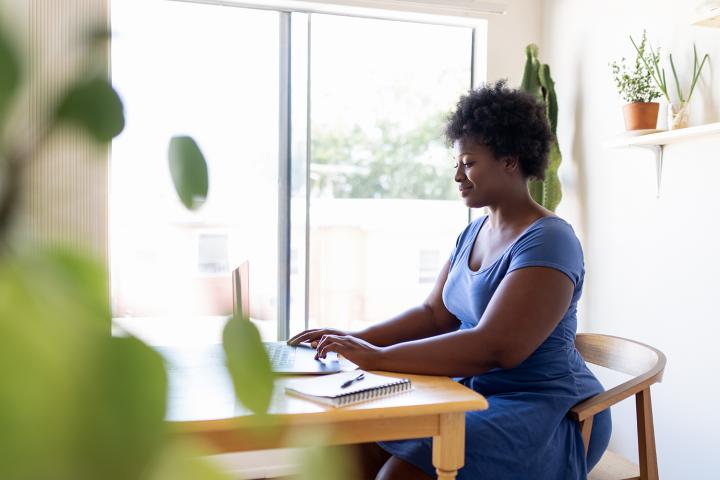 A woman sits in a sunny kitchen surrounded by plants working on a laptop