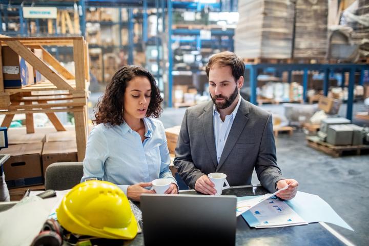 A young woman and man collaborate at a laptop on a mobile desk in a warehouse