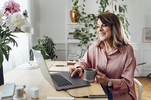 A mature woman in a pink blouse sits at her desk by a window working on her laptop.