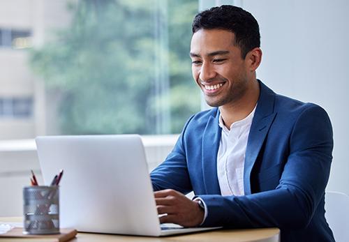 A young man of Asian Pacific heritage in a blue blazer sits at his laptop smiling and working