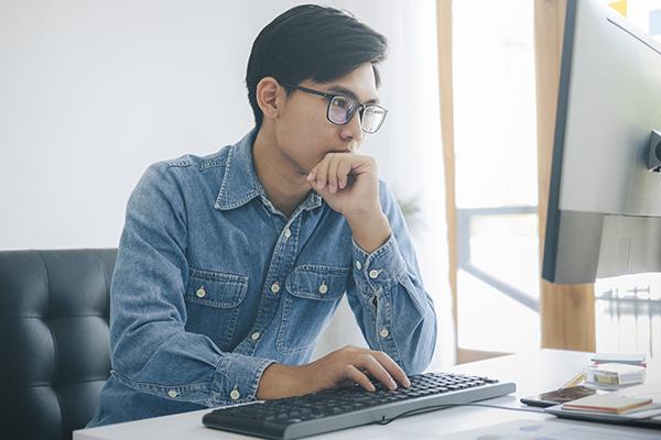 A young Asian man in a blue shirt sits at a desktop computer learning.