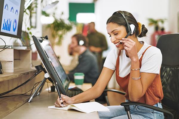 A woman in customer service sits smiling and recording information while taking a phone call. She sits in front of two computer monitors and coworkers are in the background.