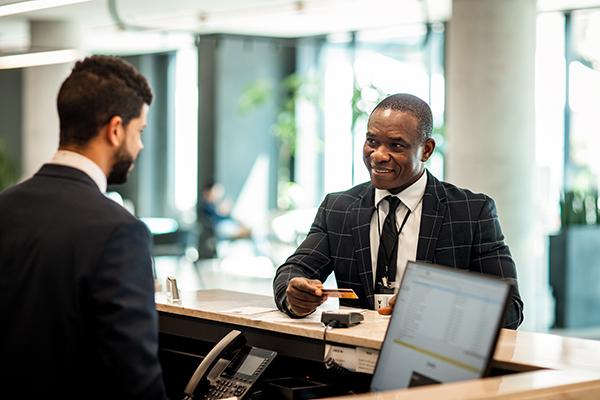 Two man stand at a hotel front desk. An African American gentleman is talking to the desk attendant, smiling, and holding a credit card.