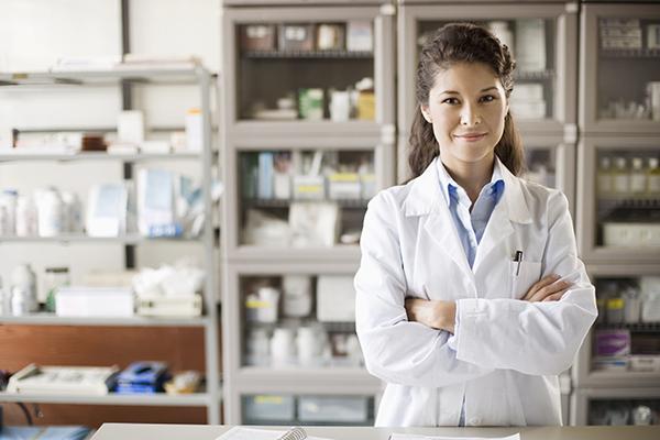 A pharmacist stands confidently in front of shelves with her arms crossed in front of her. She is smiling. 
