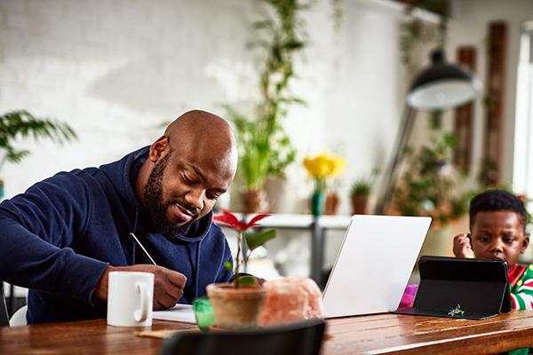 A father and son sit at a table in an airy, open room. The father is on a laptop taking a course, and the son is watching an iPad