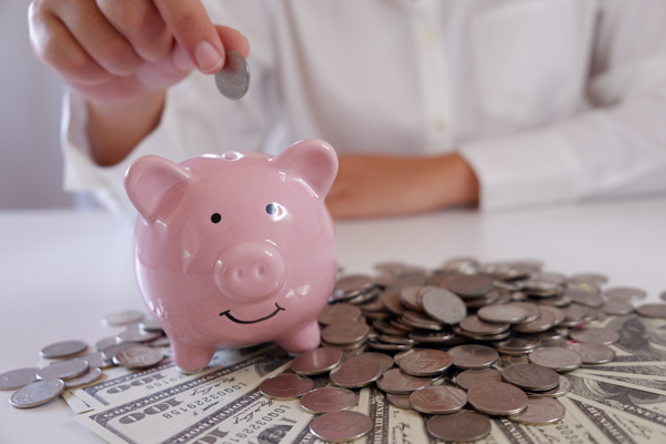 A piggy bank sits on a pile of coins and dollars while a man in a white shirt places a coin into its slot