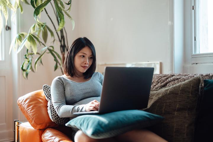 A young Asian woman works on a laptop while sitting on a couch in a living room.