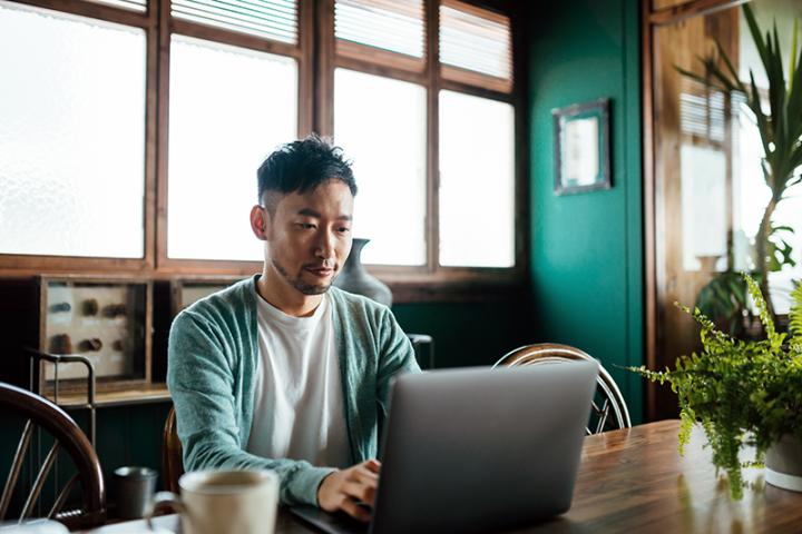 A young Asian man sits working on a laptop in a modern yet rustic kitchen.