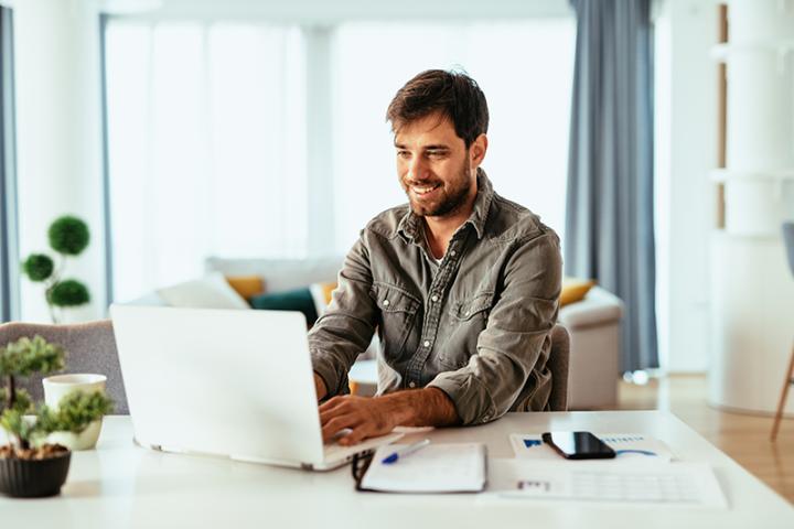 A white man with a beard sits at a white table with a white laptop smiling and learning