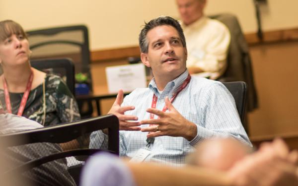 White man at a conference sitting in chair with hands gesturing as trying to accentuate a point