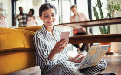 Woman sitting on floor leaning on yellow couch with a laptop in her lap and looking at a mobile phone