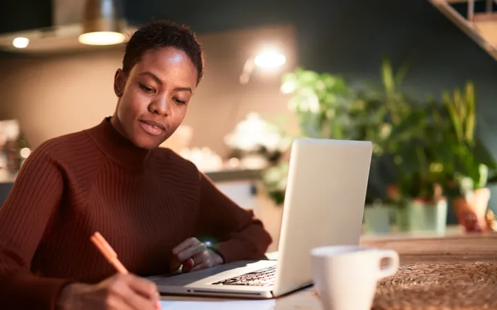 A person working at a table with a laptop and writing on paper. 