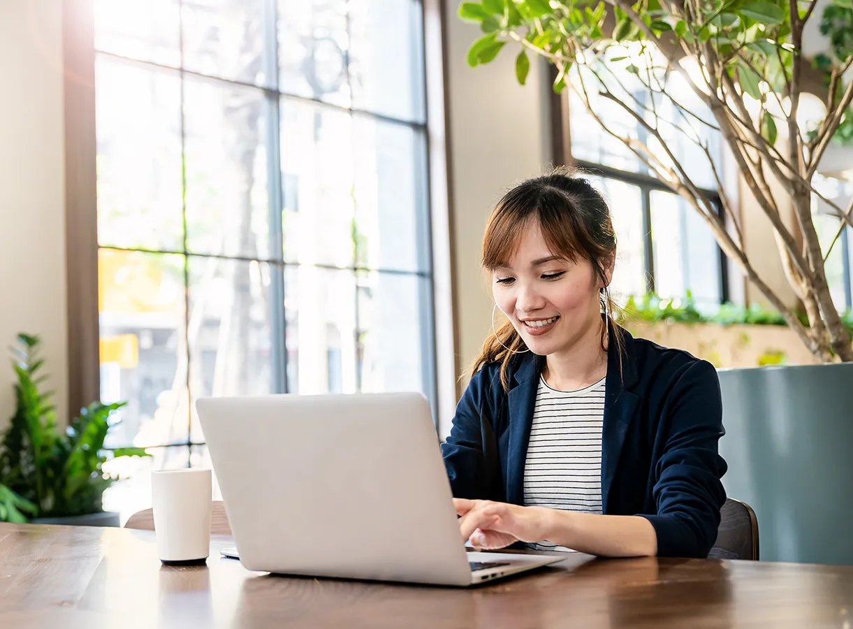 A young woman at a laptop in a sun-filled cafe and mug of coffee beside her.