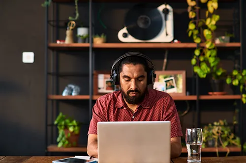 An South Asian man works on a laptop wearing headphones. Behind him is a shelf decorated with books, photos, ceramics, and plants.