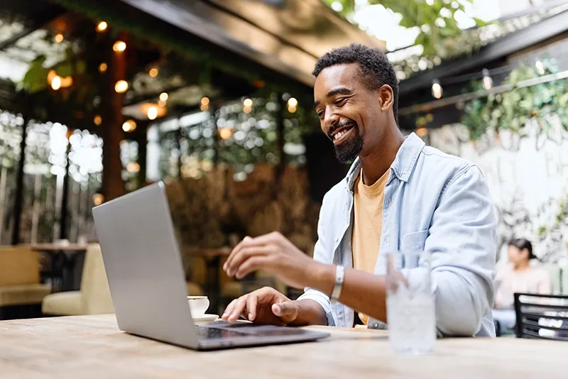 A young African American man sits in an outdoor cafe studying on his laptop and smiling