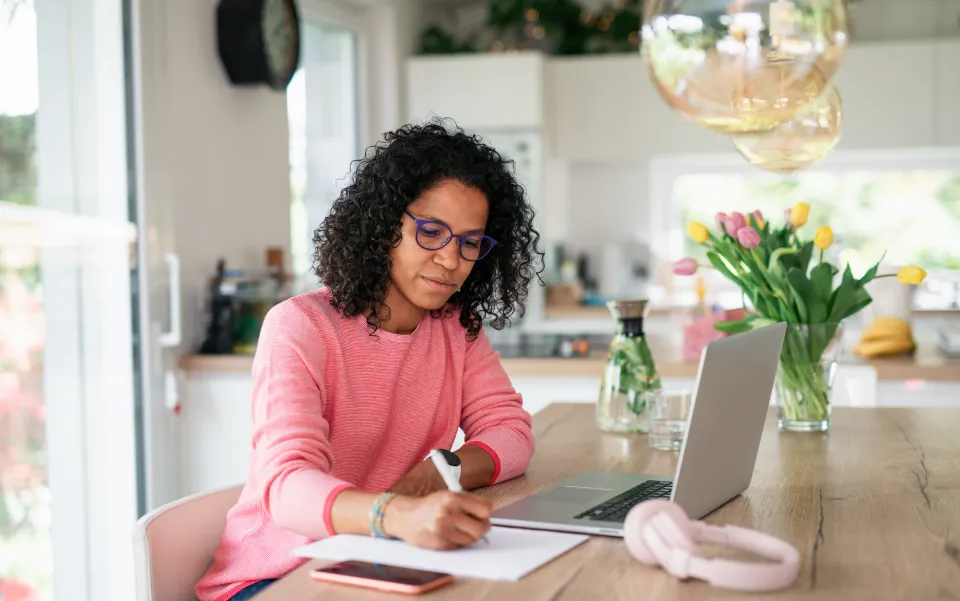 Woman wearing a pink sweater writing on a notepad in front of a laptop while sitting on a kitchen table.