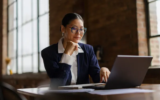 Professional woman in business attire working on a laptop at a desk in a bright, modern office.