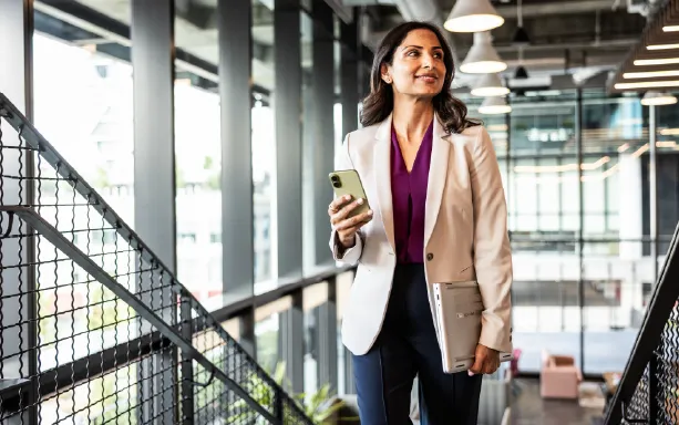 Confident businesswoman holding a phone and laptop walks through a modern office space, appearing focused and ready for the workday.